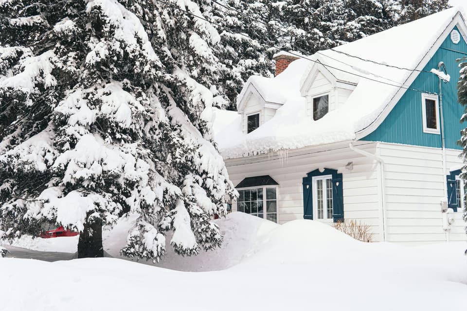 A house with dormers under deep snow, beside a snow-laden spruce
