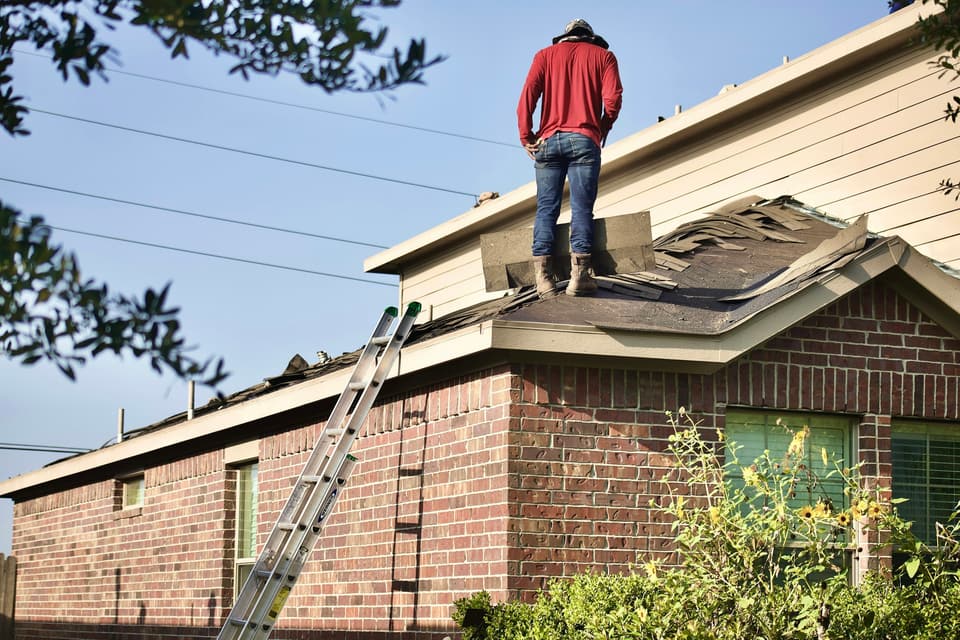 A worker standing on a brick house roof with the old shingles partly torn off and a ladder against the eave